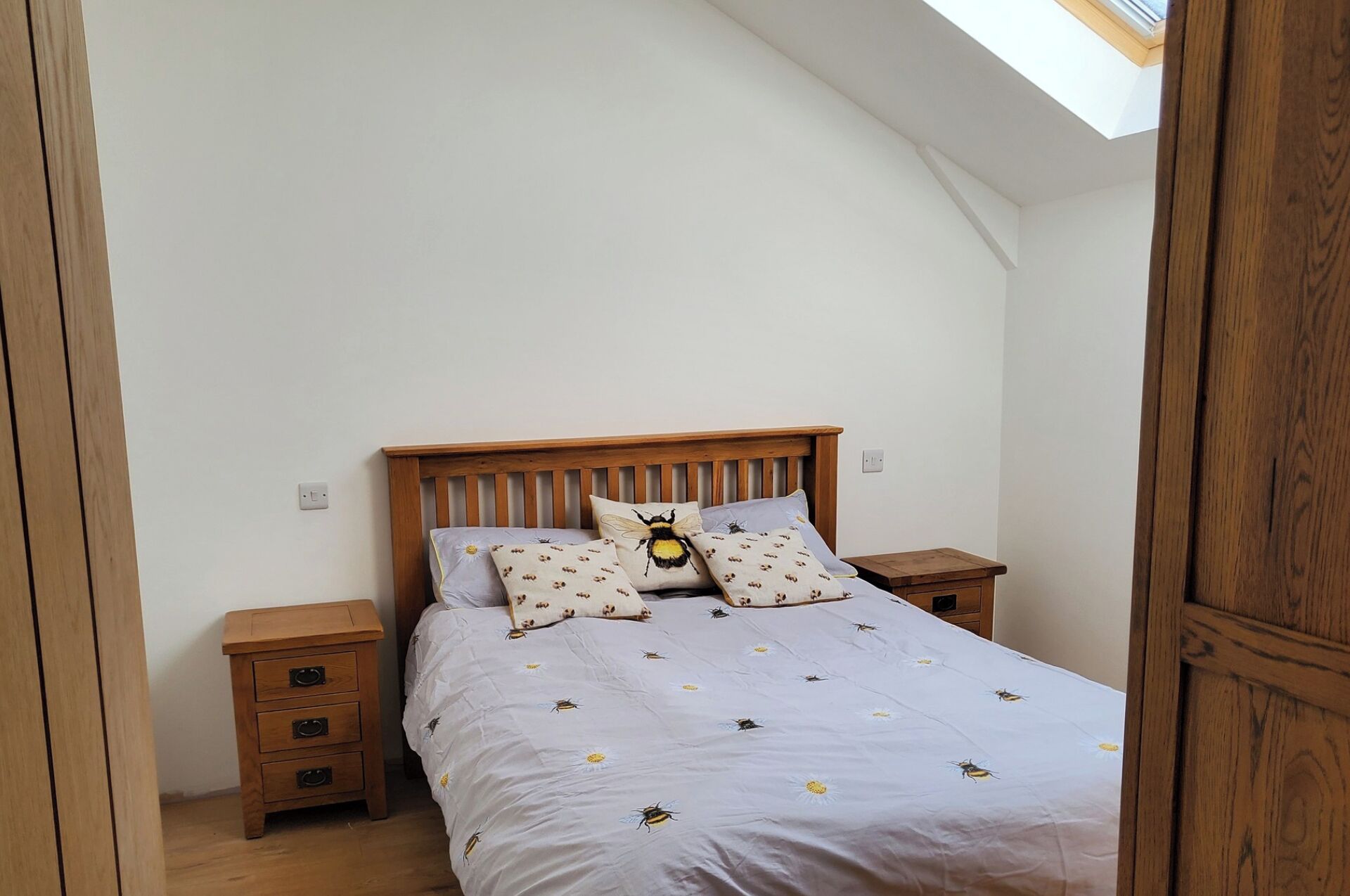 Ein Schlafzimmer mit schrägen Wänden, einem hölzernen Bettgestell, einer weißen Bettdecke mit Bienenmuster, zwei Nachttischen und einem Oberlicht.