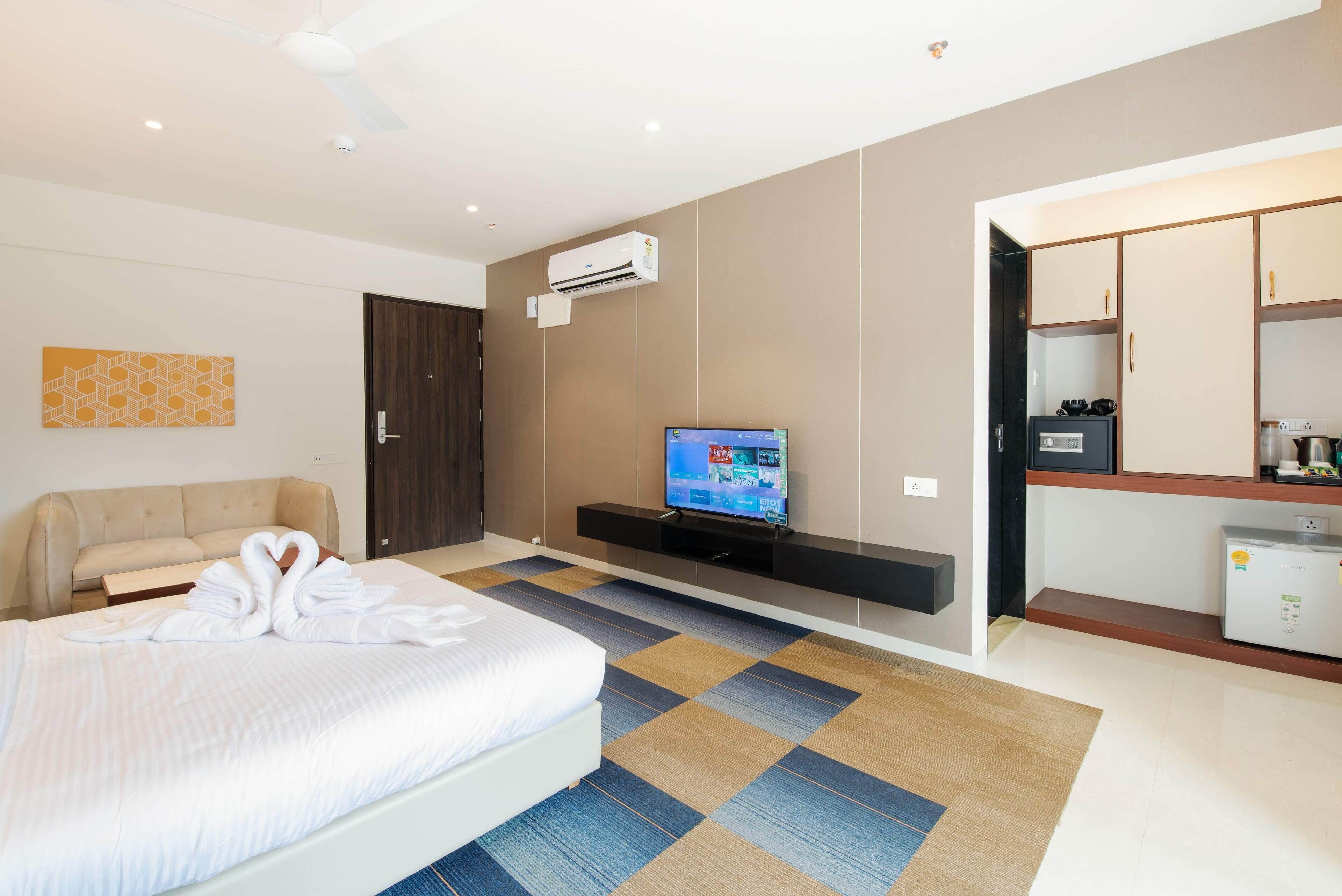 Quarto de hotel moderno com cama, televisão, sofá e cozinha compacta.
