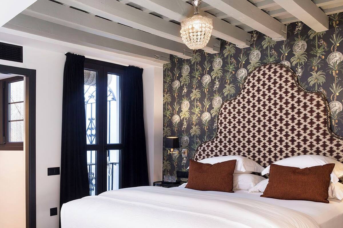 Un dormitorio con una cama grande, un banco, una araña y un papel tapiz estampado.