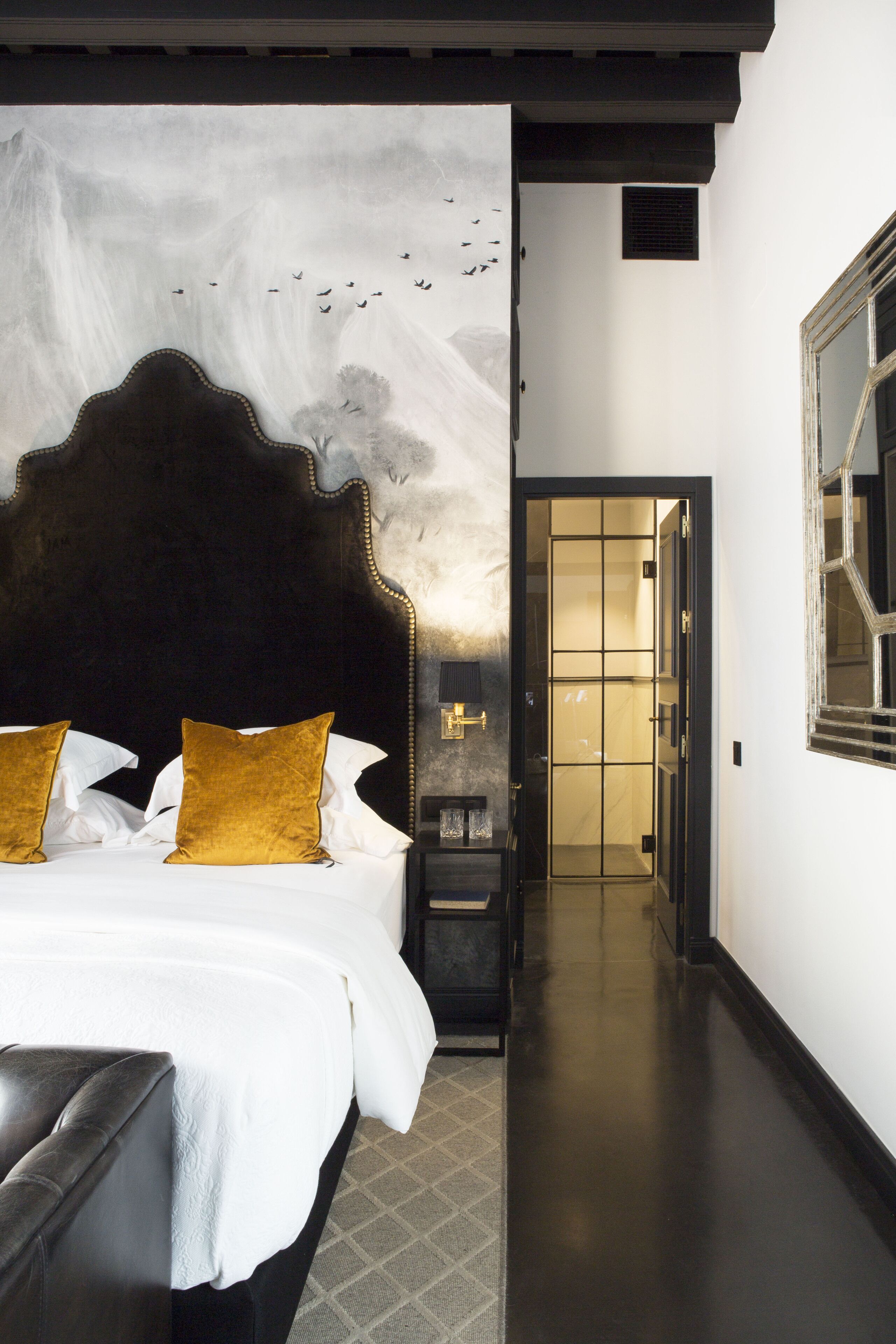A hotel room with a large bed, a headboard, a nightstand, and a wall-mounted lamp.