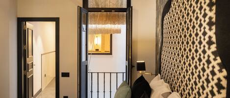 Una habitación de hotel con una cama, una mesita de noche con una lámpara, un cabecero estampado y vistas a un pasillo y otra habitación a través de una puerta abierta.