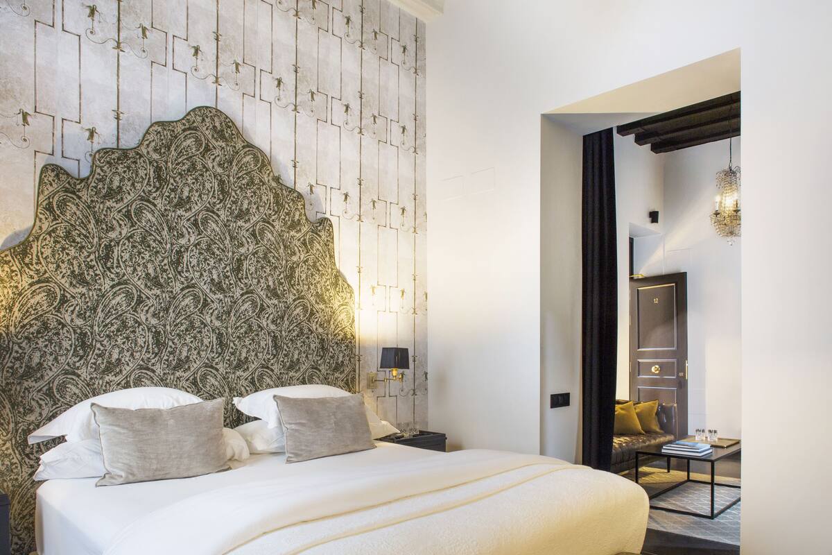 Un dormitorio con un gran cabecero decorativo, una cama con sábanas blancas, una mesita de noche con lámpara y vistas a otra habitación a través de una puerta abierta.