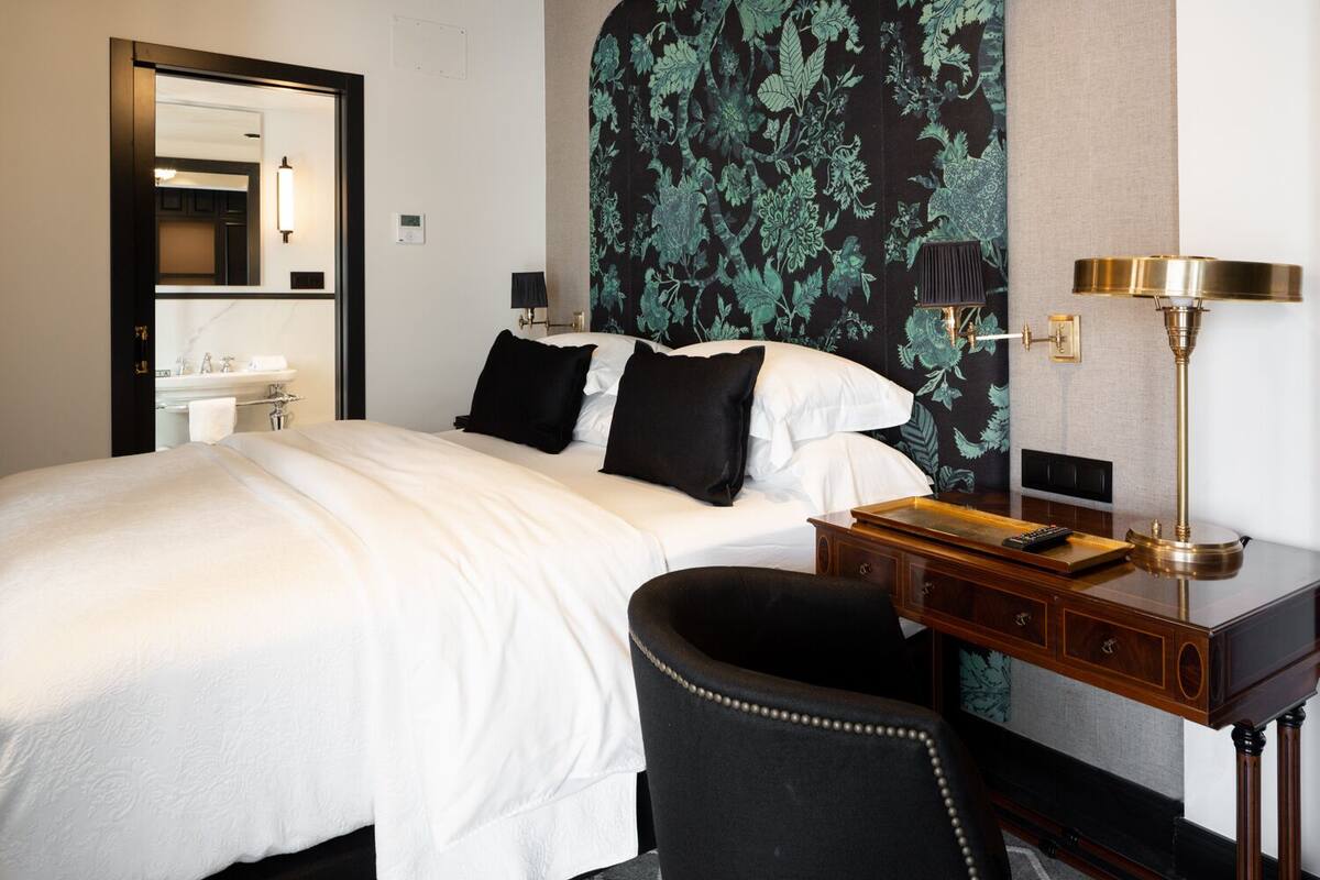 Habitación de hotel con cama, escritorio con lámpara, silla y baño visible a través de una puerta abierta.