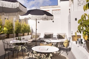 Un patio con mobiliario exterior, incluyendo mesas y sillas, rodeado de plantas en macetas y sombrillas.