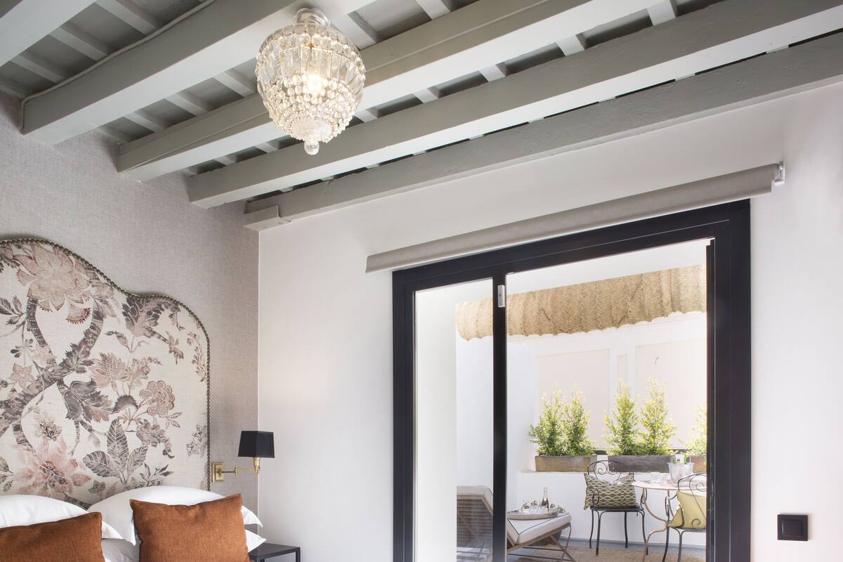 Un dormitorio con una cama grande, una araña, una mesita de noche, una lámpara y vistas a un patio exterior con mesa y sillas.