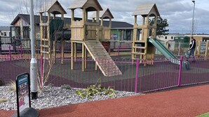 Ein Spielplatz mit hölzernen Klettergerüsten und Rutschen, umgeben von einem violetten Zaun.