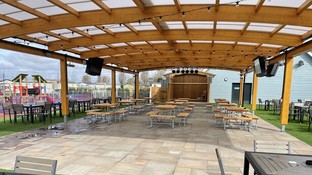 Eine überdachte Terrasse im Freien mit Holztischen und -stühlen, eine Bühne mit Lautsprechern und ein Spielbereich für Kinder im Hintergrund.
