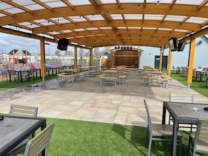 Eine überdachte Terrasse im Freien mit Holztischen und -stühlen, eine Bühne mit Lautsprechern und ein Spielbereich für Kinder im Hintergrund.