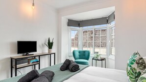 A modern hotel room with a bed, a desk with a computer, a chair, and a large window with a view of buildings. - Ivydene - Beautiful Cottage Stone Throw From Beach Wonderful Sea Views (Saundersfoot)