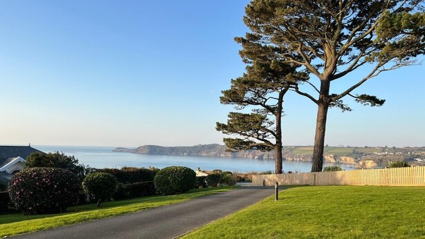 A residential street with a paved path, a large tree, and a view of the sea and hills. - Beautiful Sea view apartment in Carlyon Bay (Carlyon Bay)