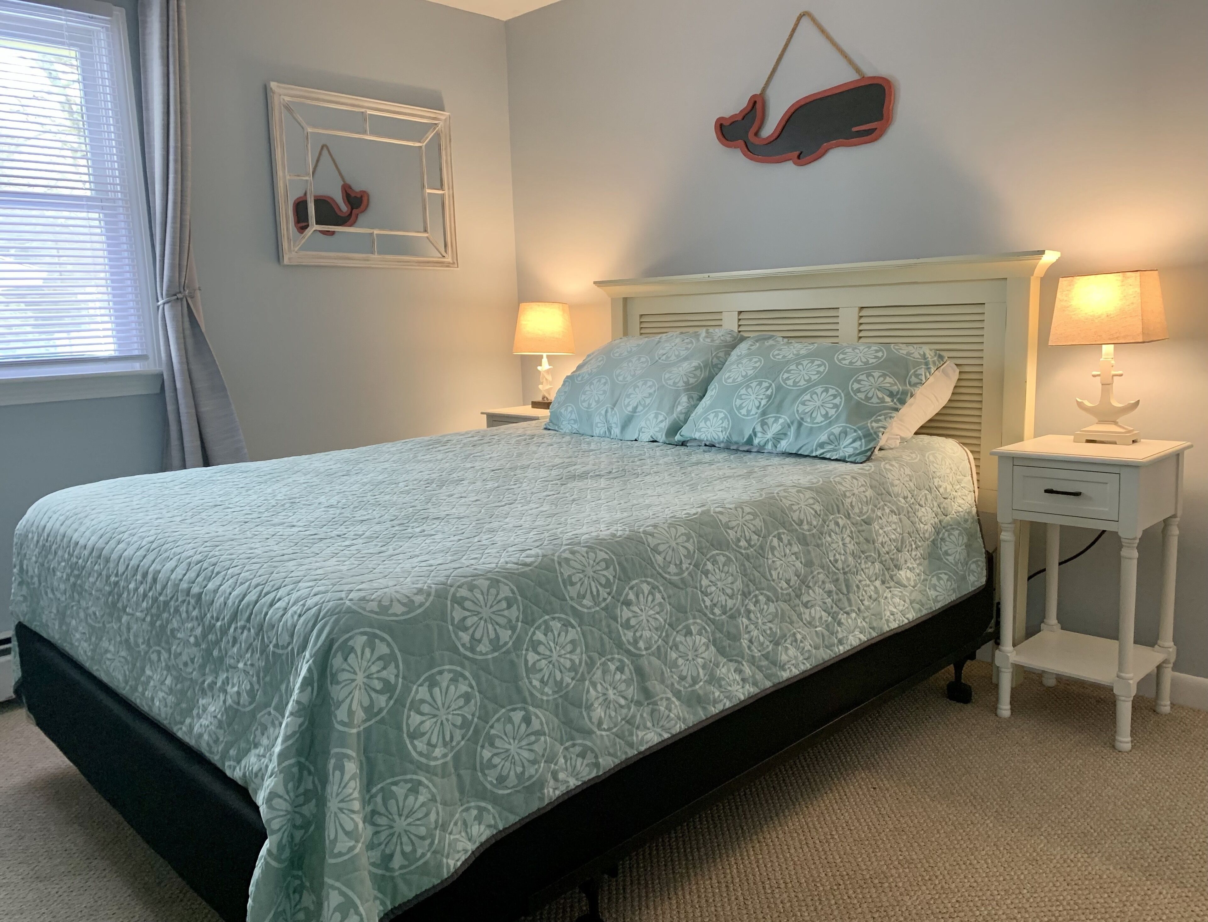 Une chambre à coucher avec un lit, deux tables de chevet avec des lampes, une décoration murale représentant une baleine et un tableau encadré.