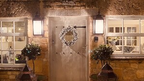 A stone building with a wooden door, two lanterns, and a wreath on the door. - Stunning Interior designed cottage (Chipping Campden)