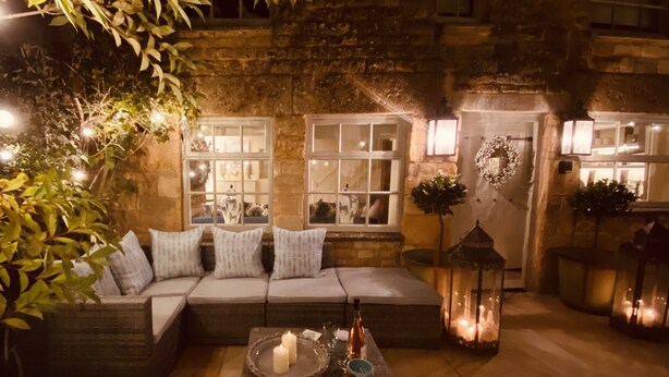 An outdoor seating area with a sofa, cushions, and a table with candles and a bottle. - Stunning Interior designed cottage (Chipping Campden)