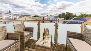 Una terraza en la azotea con muebles de mimbre, una mesa con una botella y vasos, y vistas de edificios circundantes.