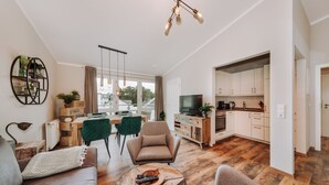 Una sala de estar moderna con un sofá, una mesa de centro y un televisor. Se observa un comedor con una mesa y sillas. Se puede ver la cocina, con armarios y una encimera.