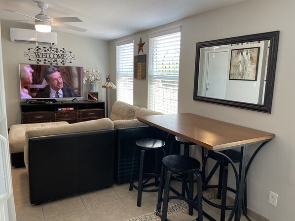 Una sala de estar moderna con un sofá seccional, una mesa de comedor de madera con taburetes altos, un televisor y decoración en la pared.