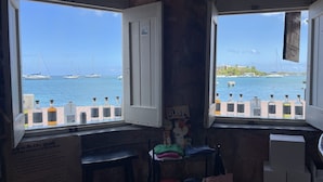 Een kamer met twee ramen met uitzicht op een jachthaven vol zeilboten. Er staat een klein tafeltje met een stoel en wat spullen op.