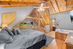 A bedroom with a sloped ceiling, a bed with grey bedding, a wooden headboard, a window, and a view of the outdoors.
