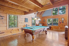 A wooden interior with a pool table, large windows, and wooden beams.