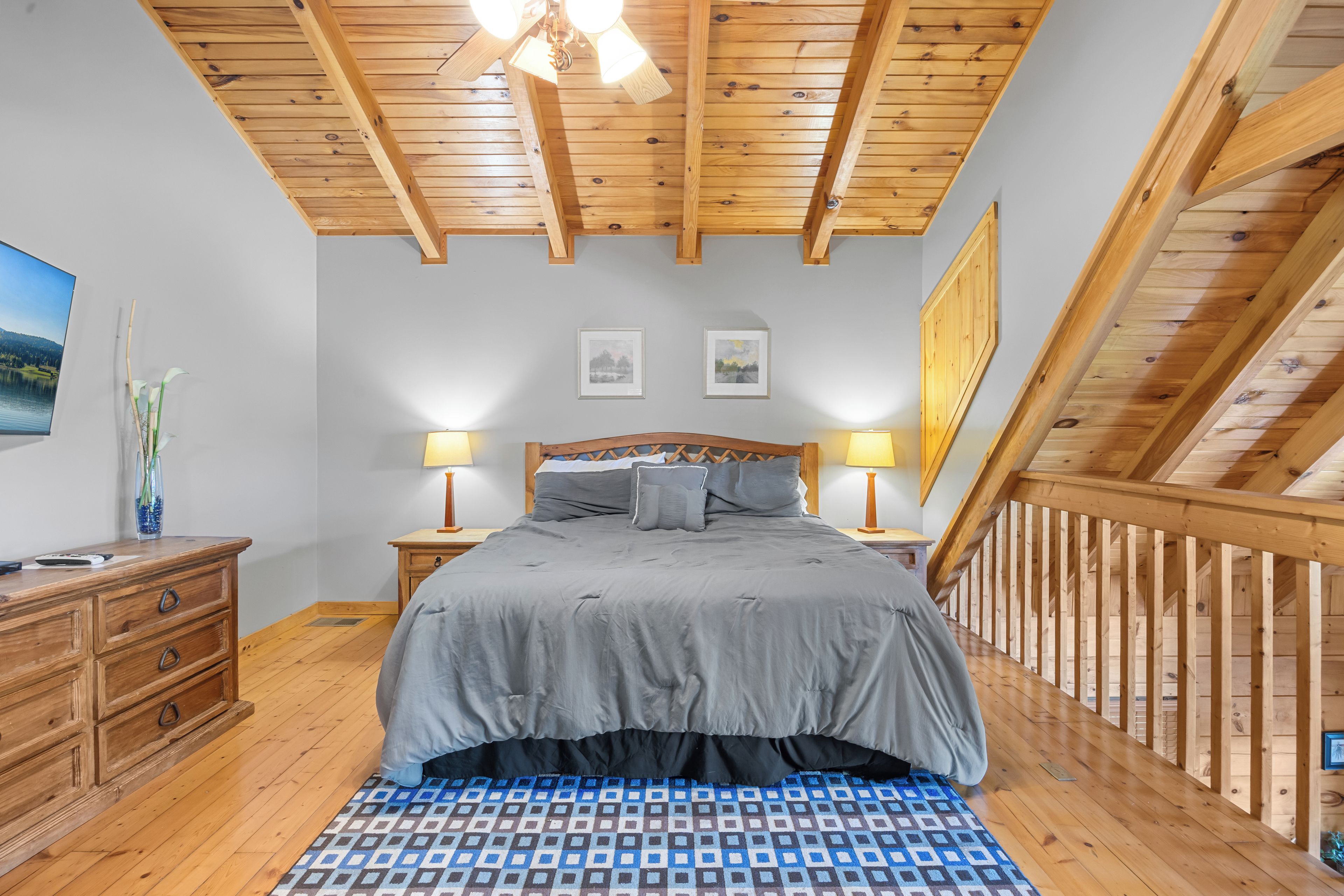 A bedroom with a wooden ceiling, a bed with gray bedding, two bedside lamps, a nightstand, a dresser, and a framed picture on the wall.