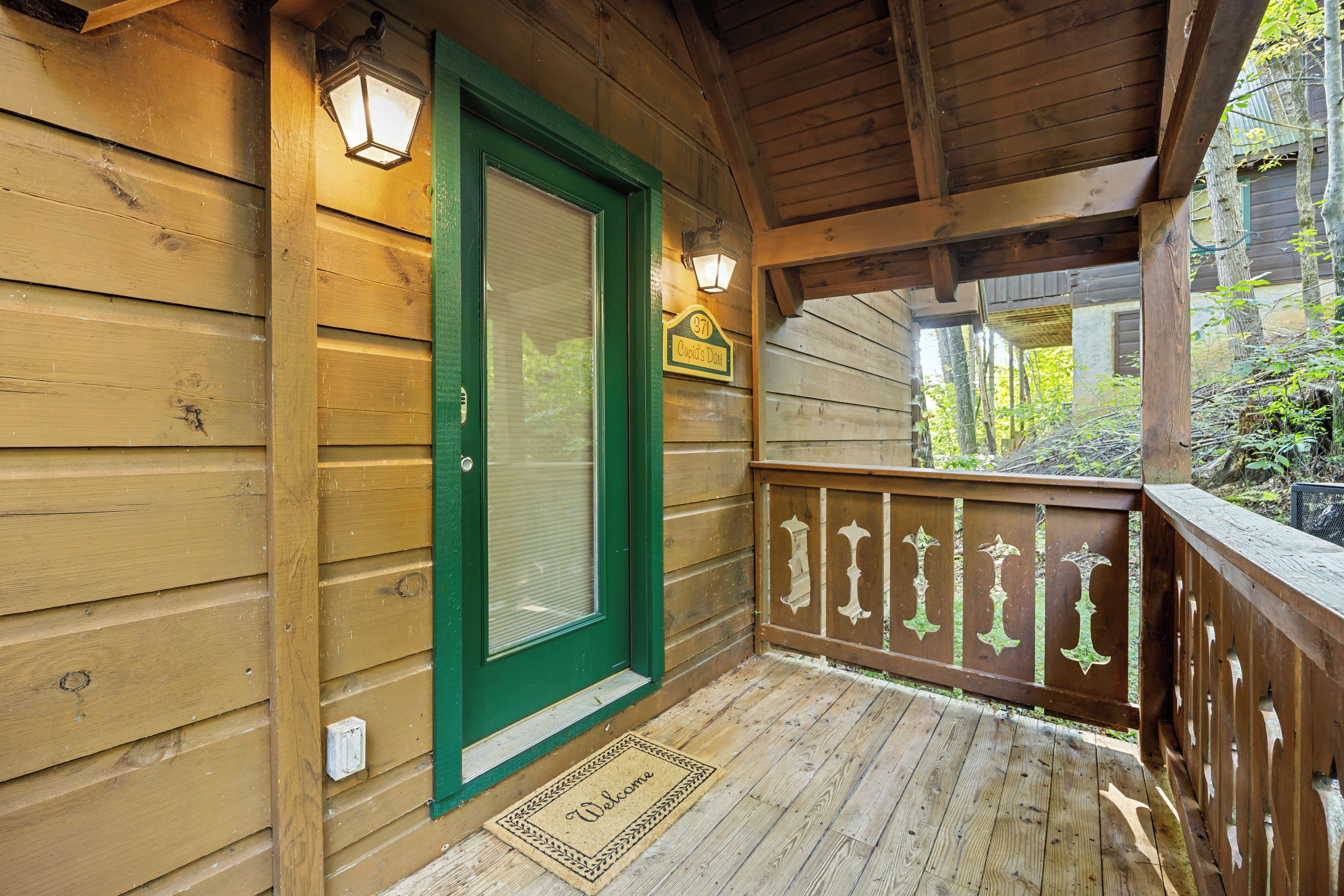 A wooden porch with a green door, a welcome mat, and a sign indicating the property number.