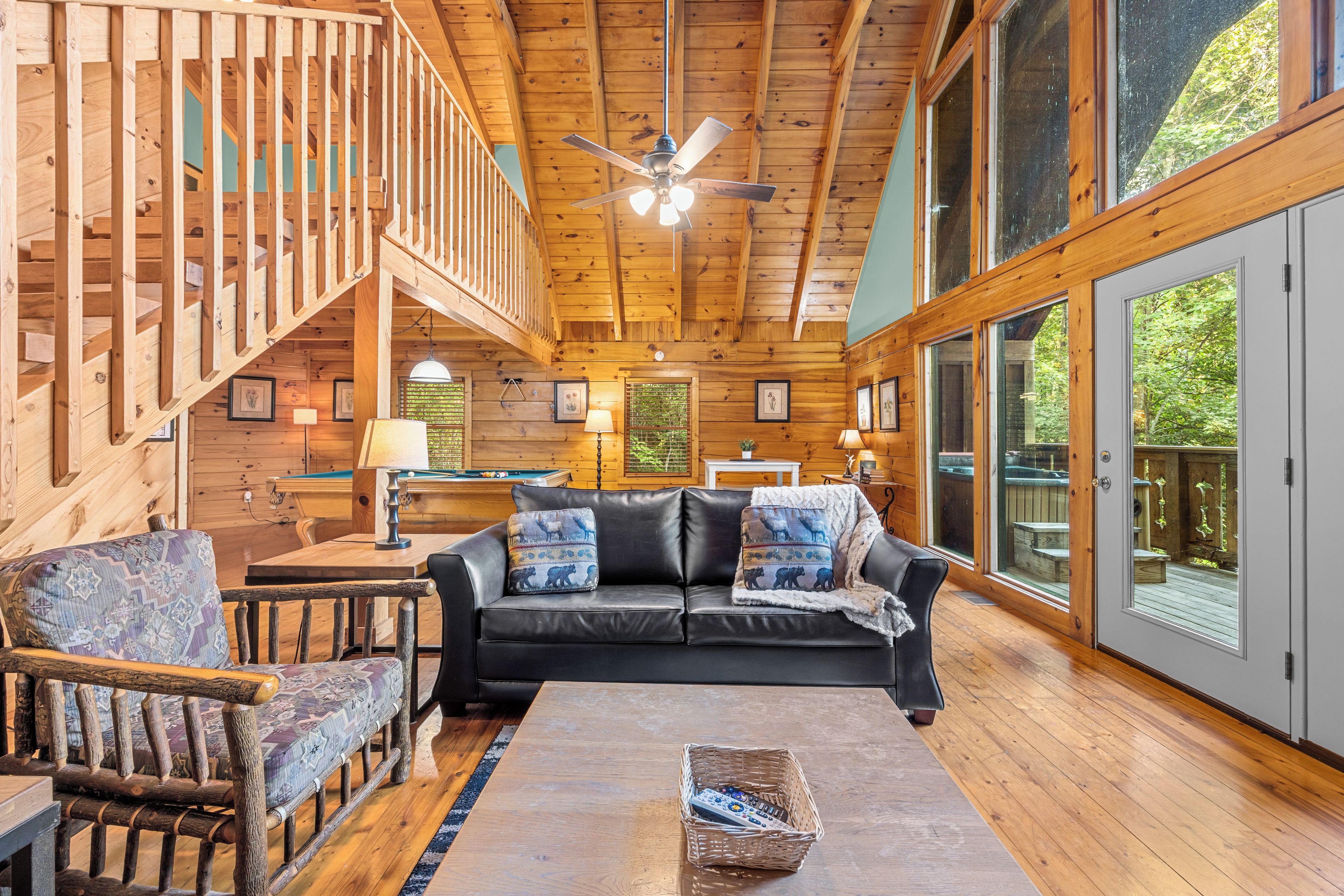 A cozy cabin interior with a wooden staircase, a black leather sofa, and a pool table.
