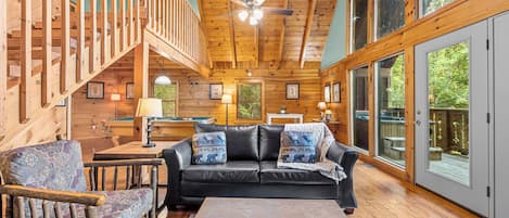 A cozy cabin interior with a wooden staircase, a black leather sofa, and a pool table.