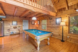 A wooden interior with a pool table, a refrigerator, and framed pictures on the walls.