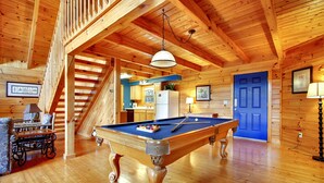 A wooden interior with a pool table, a staircase, and a kitchen area.