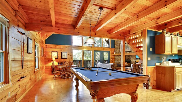 A wooden interior with a pool table, wooden cabinets, and large windows.