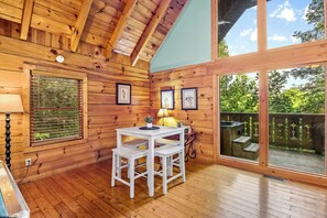 A wooden cabin interior with a dining table, benches, framed pictures, and a hot tub outside.