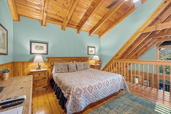 Un dormitorio con techo de madera, una cama con ropa de cama estampada, dos lámparas de noche, una mesita de noche con una lámpara, un cuadro enmarcado y una ventana con vistas.