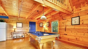 A wooden cabin interior with a pool table, a refrigerator, and a window with a view of greenery.