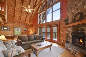 A cozy cabin interior with a wooden ceiling, a stone fireplace, a sofa, a coffee table, and a pool table.