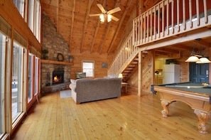 A cozy cabin interior with a fireplace, a sofa, a pool table, and a balcony.