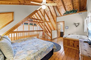 A bedroom with a sloped ceiling, a bed with a floral bedspread, a wooden dresser, and a bathroom with a bathtub.
