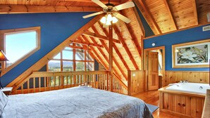 A bedroom with a large bed, a wooden ceiling, a window, a painting, and a bathroom with a bathtub. - Mystic- Situated in a very private wooded setting with great mountain views!! (Gatlinburg)