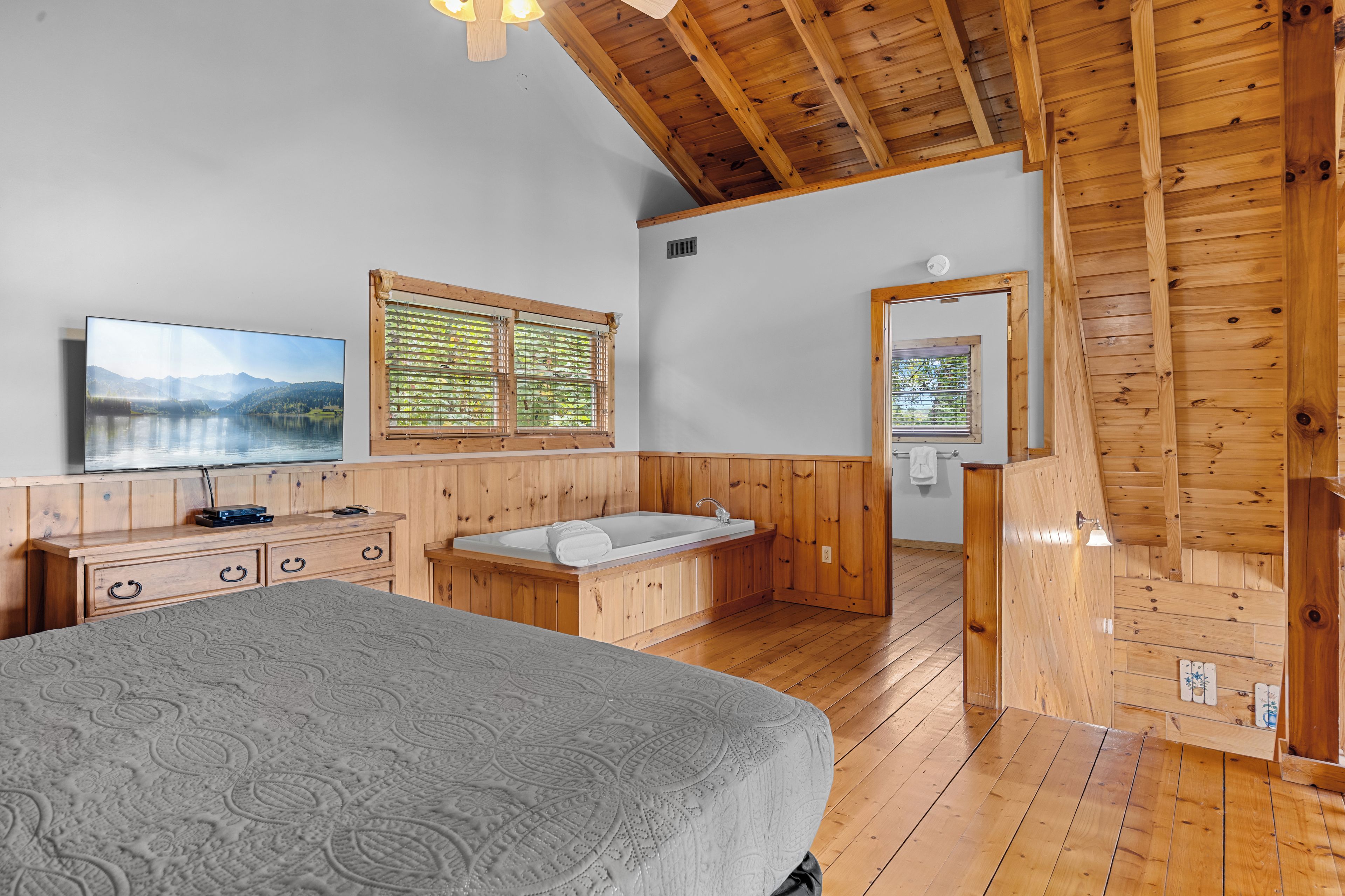 Ein Schlafzimmer einer Holzhütte mit einem Bett, einer Holzkommode, einem Fernseher und einem Fenster mit Jalousien.