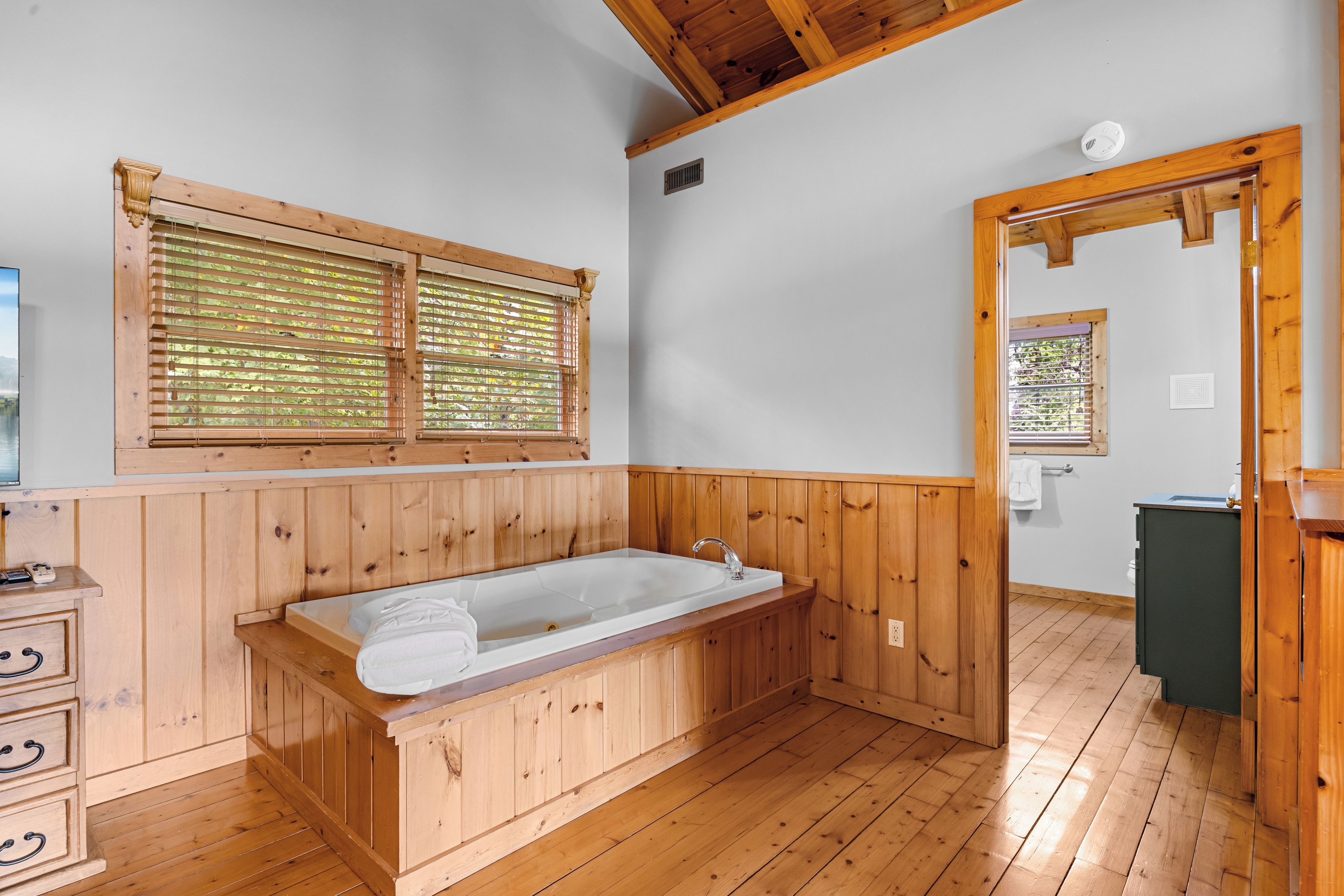 A wooden-floored bathroom with a freestanding bathtub, a window with blinds, and a wooden door.