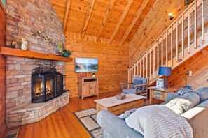 A cozy cabin interior with a fireplace, wooden walls, a sofa, and a staircase.