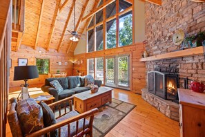 Interior acogedor de una cabaña con techo de madera, chimenea de piedra y un ventanal con vistas a la vegetación.