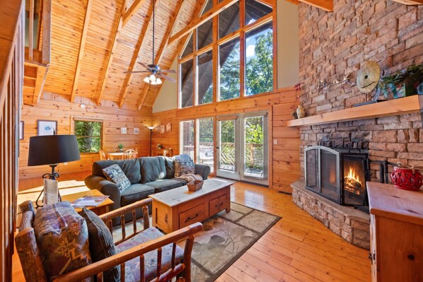 A cozy cabin interior with a wooden ceiling, a stone fireplace, and a large window overlooking greenery.