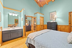 A bedroom with a large bed, a dresser, a vanity with a mirror, and a door leading to another room.