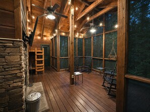 Interior de una cabaña de madera con pared de piedra, suelo de tablas y ventilador de techo.