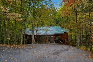 Een chalet met een metalen dak, omringd door bomen met herfstbladeren, en een grindpad dat naar de ingang leidt.