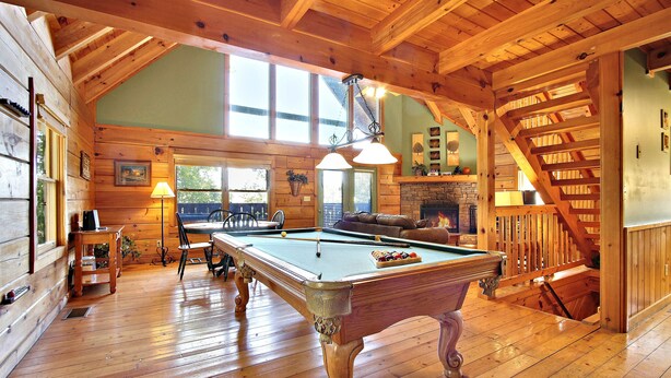 A wooden pool table in a room with high ceilings and wooden beams, a fireplace, and a staircase.