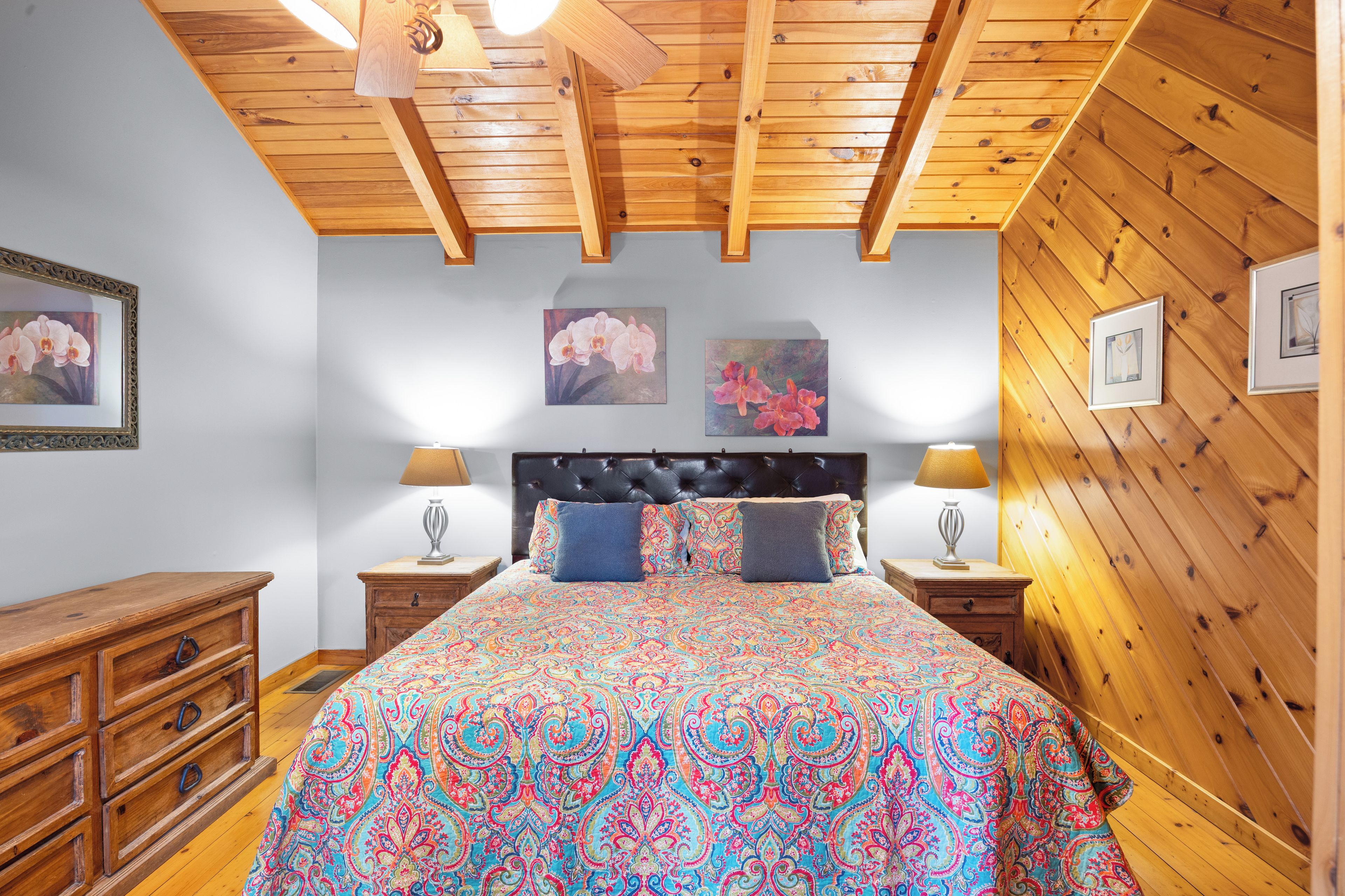 A bedroom with a wooden ceiling, a bed with patterned bedding, two nightstands with lamps, a dresser, and framed artwork on the walls.