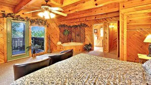 A wooden cabin bedroom with a bed, a ceiling fan, a window with blinds, a small table, and a door leading to another room.