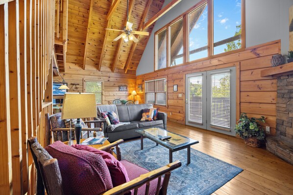 A cozy cabin interior with a wooden ceiling, a fireplace, a sofa, a coffee table, and a large window.