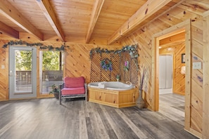 A wooden cabin interior with a wooden floor, a red chair, a wooden bathtub, and a door leading to another room.